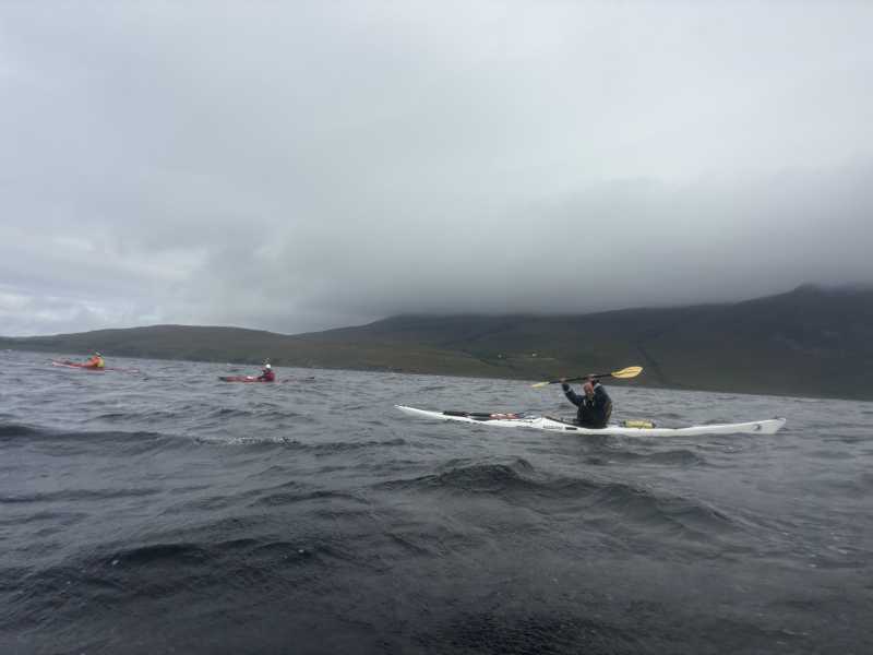 Three paddlers on grey choppy water, low cloud sitting on the hills behind